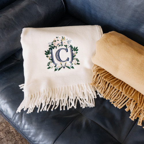 Custom embroidered wedding crest on blanket. Cream and camel colored blankets with floral embroidery in pink, cream and green thread. Monogram in navy thread. Shown on a dark couch.