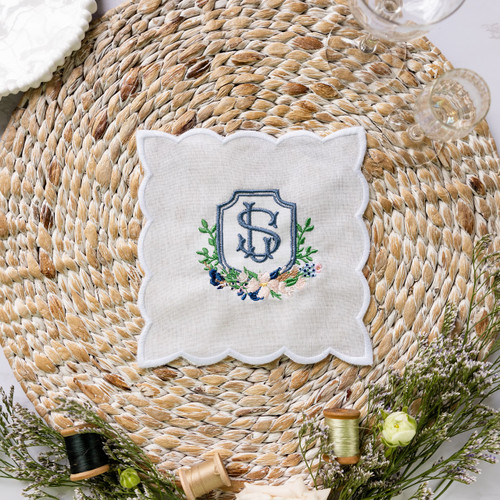 Custom embroidered white scalloped edge linen cocktail napkin. Embroidery is of the couple's wedding crest in smokey blue, navy, pink and green thread. Cocktail napkin shown on woven placemat with flowers and threads as decoration.