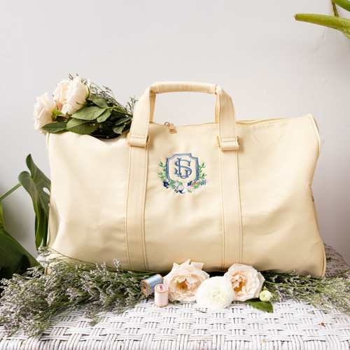 Custom embroidered duffle bag personalized with the couple's wedding crest. Embroidered in smokey blue, pink, green and navy thread. Shown on a table with flowers for decoration. 