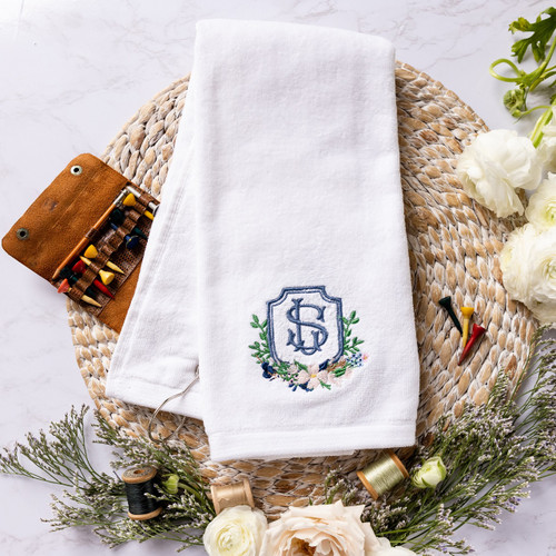 Custom embroidered golf towel for bride or groom. White gold towel is embroidered with their wedding crest. Shown with golf tees and flowers on a woven mat for decoration.