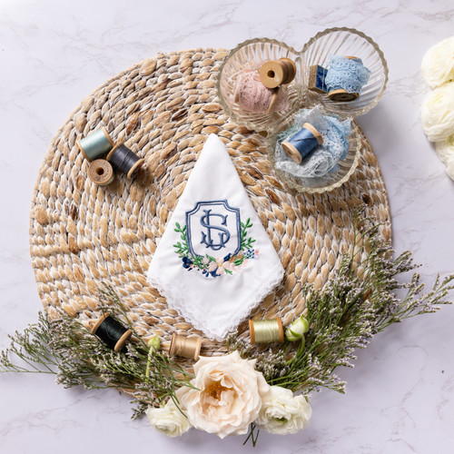 Custom embroidered handkerchief for the bride, parents of the bride using the couple's wedding crest. Personalized embroidery in smokey blue, navy, pink and green. Shown on woven mat with flowers and embroidery thread for decoration.
