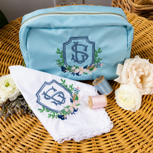 Our happy tear custom embroidered package for the bride on her wedding day. Items that are embroidered are handmade handkerchief and cosmetic bag. All are personalized with the bride's wedding crest. Wedding crest embroidery is done in smokey blue, navy, green and pink threads. Shown on a woven table with flowers and cosmetic items for decoration.