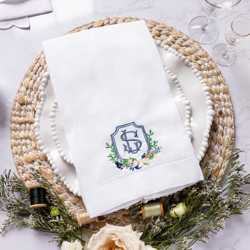 Custom embroidered tea towel using couple's wedding crest. Embroidered in smokey blue, navy, pink and green thread. White linen tea towel shown on white plates with flowers and thread as decoration.