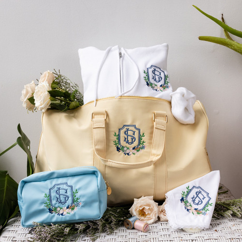Our luxury custom embroidered package for the bride on her wedding day. Items that are embroidered are handmade handkerchief, cosmetic bag, duffle bag and zip up hoodie. All are personalized with the bride's wedding crest. Wedding crest embroidery is done in smokey blue, navy, green and pink threads. Cosmetic bag is shown on a woven table with flowers and cosmetic items for decoration.