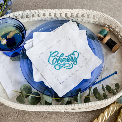 Custom embroidered cocktail napkins. Set of 4. Embroidered with a "cheers" in turquoise thread. Background has a basket, thread, flowers, glass and a spoon.