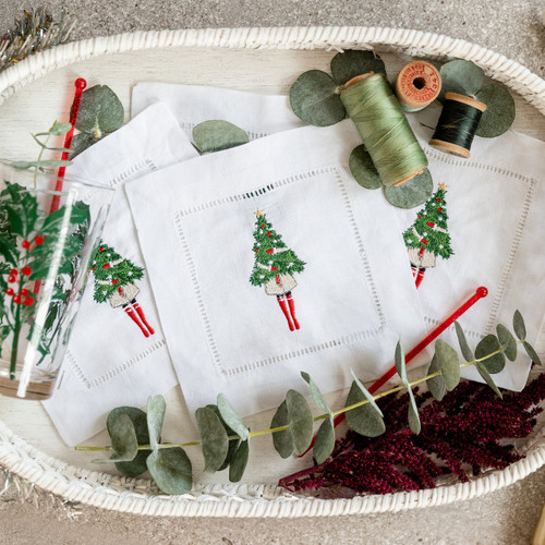 A set of 4 cocktail napkins embroidered with a woman holding a Christmas tree. The background has thread, flowers, a glass and stirrers in a basket.