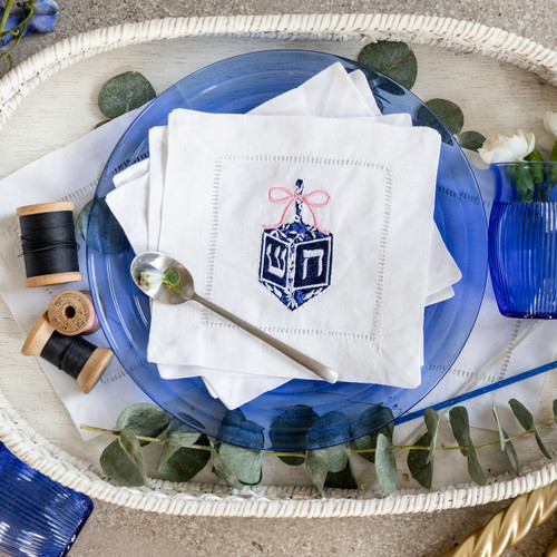 Custom embroidered cocktail napkins. Set of 4. Embroidered with a dreidel in blue, white and pink thread. Background has a basket, flowers, thread, glasses and a spoon.