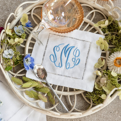 Set of 4 cocktail napkins embroidered with a three letter monogram in turquoise thread. The background has a basket, flowers, a spoon and a glass.