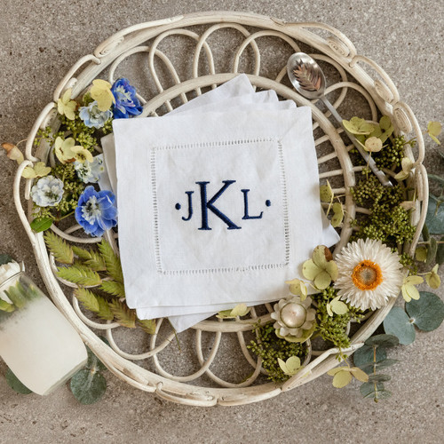 Set of 4 cocktail napkins embroidered with a three letter monogram in navy thread. The background has a basket, flowers, a spoon and a glass.
