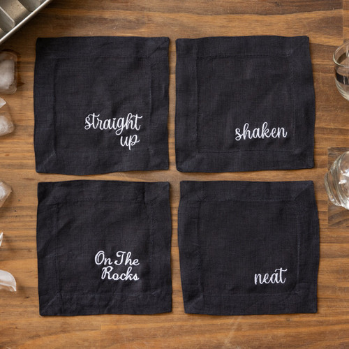 4 black cocktail napkins from Philadelphia Wedding Magazine embroidered with Straight Up, Shaken, Neat, On the Rocks in white thread. Shown on wood table with ice cubes and drinking glasses.