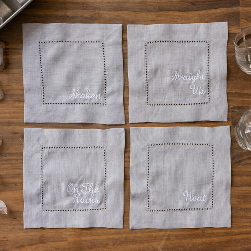 4 grey gray cocktail napkins from Philadelphia Wedding Magazine embroidered with Straight Up, Shaken, Neat, On the Rocks in white thread. Shown on wood table with ice cubes and drinking glasses.