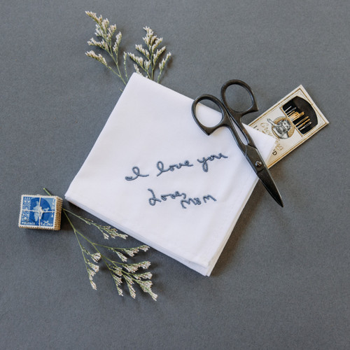 Handwriting embroidered on a white handkerchief in smokey blue embroidery thread. Pictured with sewing accessories , flowers and stamps.