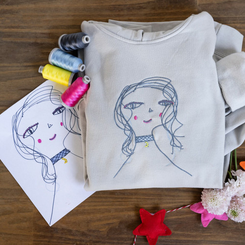 Gray hoodie sweatshirt embroidered with kid's drawing in blue, pink and yellow. Shown with embroidery threads and craft decorations.