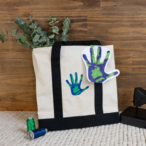 Custom embroidered tote bag personalized with your child's drawing. Drawing embroidery is done in blue and green threads. Bag is shown on a woven matte with wood grain background. There are flowers and embroidery threads.