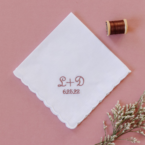 Scalloped edge white handkerchief with monogram and wedding date in dusty rose thread color on pink background.
