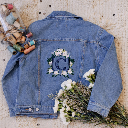 Custom embroidered wedding crest on jean jacket. Floral embroidered crest in pink, cream and green thread. Monogram in navy thread. Shown on cream color material, flowers and embroidery threads for decoration.