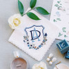 White lace handkerchief embroidered with a custom crest designed by Papergarten. Crest is blue with flowers, butterfly and other delicate flourishes.